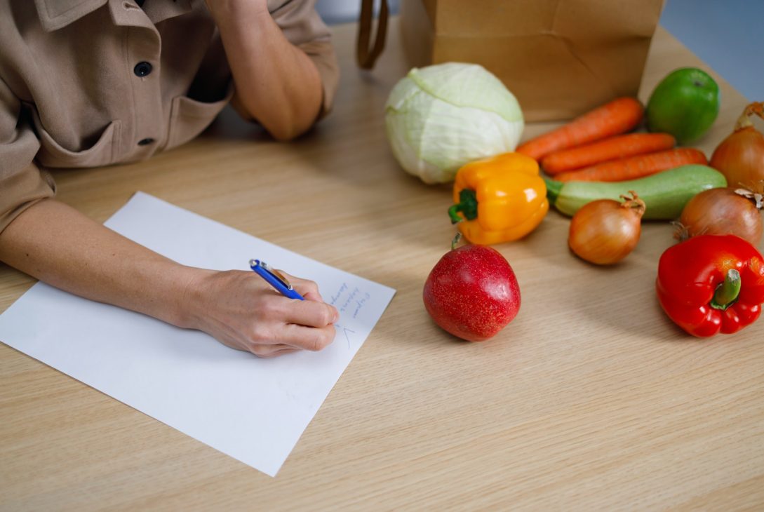 Close up view of woman writing shopping list or cooking recipe on table with many vegetables