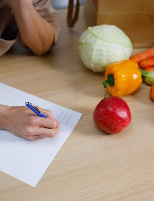 Close up view of woman writing shopping list or cooking recipe on table with many vegetables