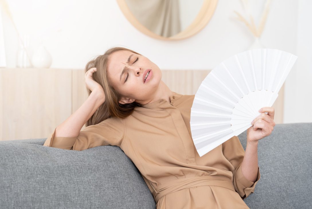 Girl on gray couch sitting at home experiencing impact of hot summer temperatures, fanning